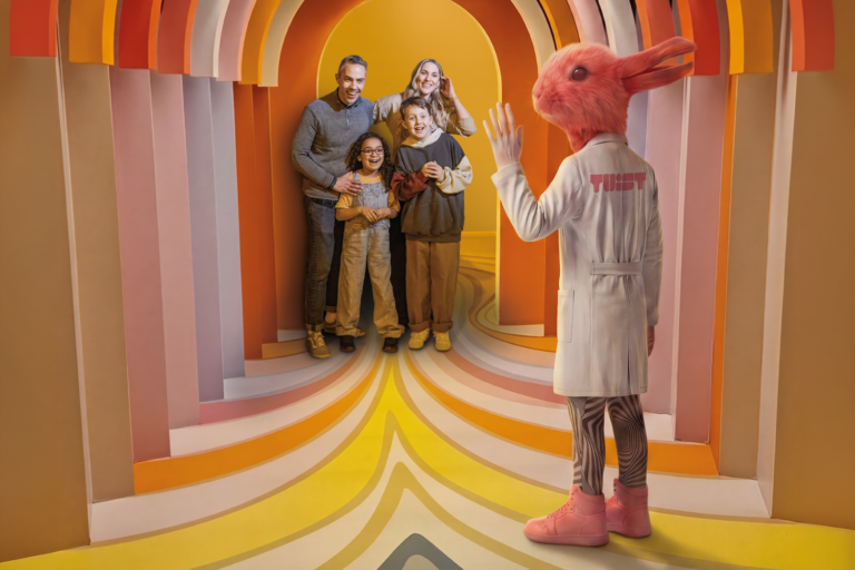 Family of four poses for a photo with a pink rabbit mascot in a white coat inside a colorful rainbow tunnel who greets them.